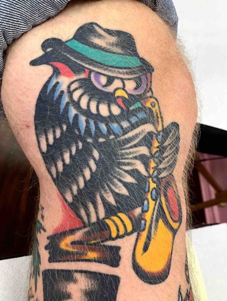 Eule mit Hut spielt Saxophon, bunte Tätowierung auf dem Oberarm.