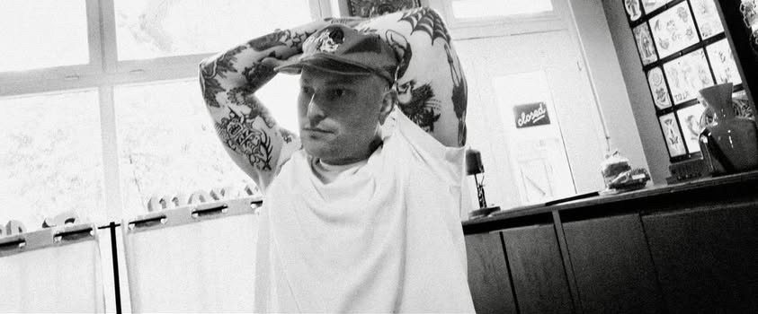 Traditional tattoo Berlin – Tattoos by Mariano – Mariano mit tätowierten Armen und Kappe, der entspannt in einem hellen Raum posiert.