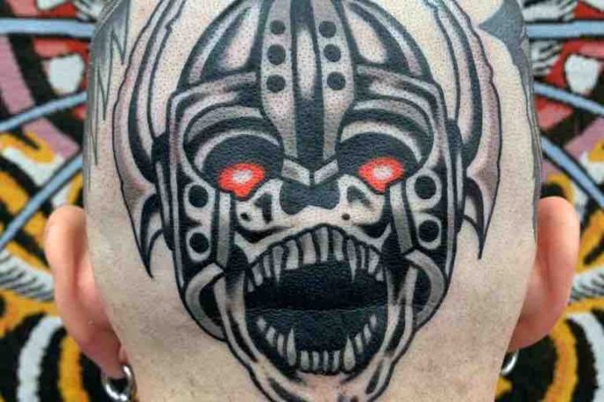 Tattoo eines grimmigen Gesichts mit roten Augen auf einer Glatze.